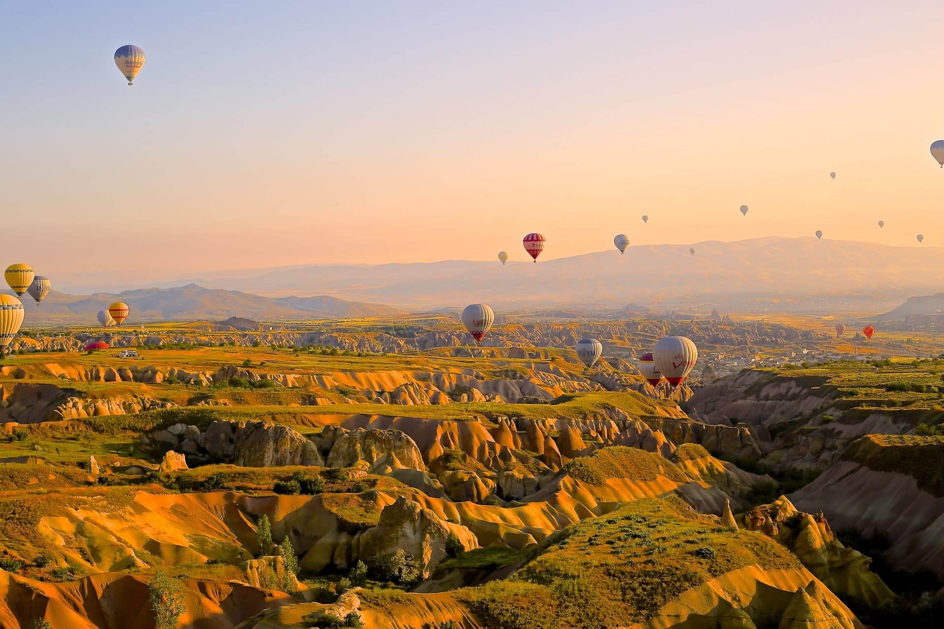 Air balloons in Turkey on a Jewish heritage tour of Turkey. They rise above green mountaintops and cliffs, illuminated by a bright orange sky,
